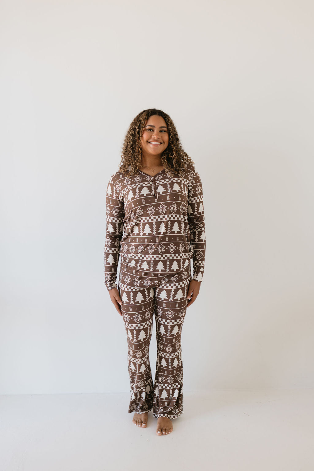 A person with curly hair is smiling while wearing the Women's Bamboo Pajamas from forever french baby, crafted to be hypo-allergenic. The pajamas feature a festive holiday pattern of trees and snowflakes against a cozy brown fabric, as they stand against a plain white background, radiating cozy holiday cheer.