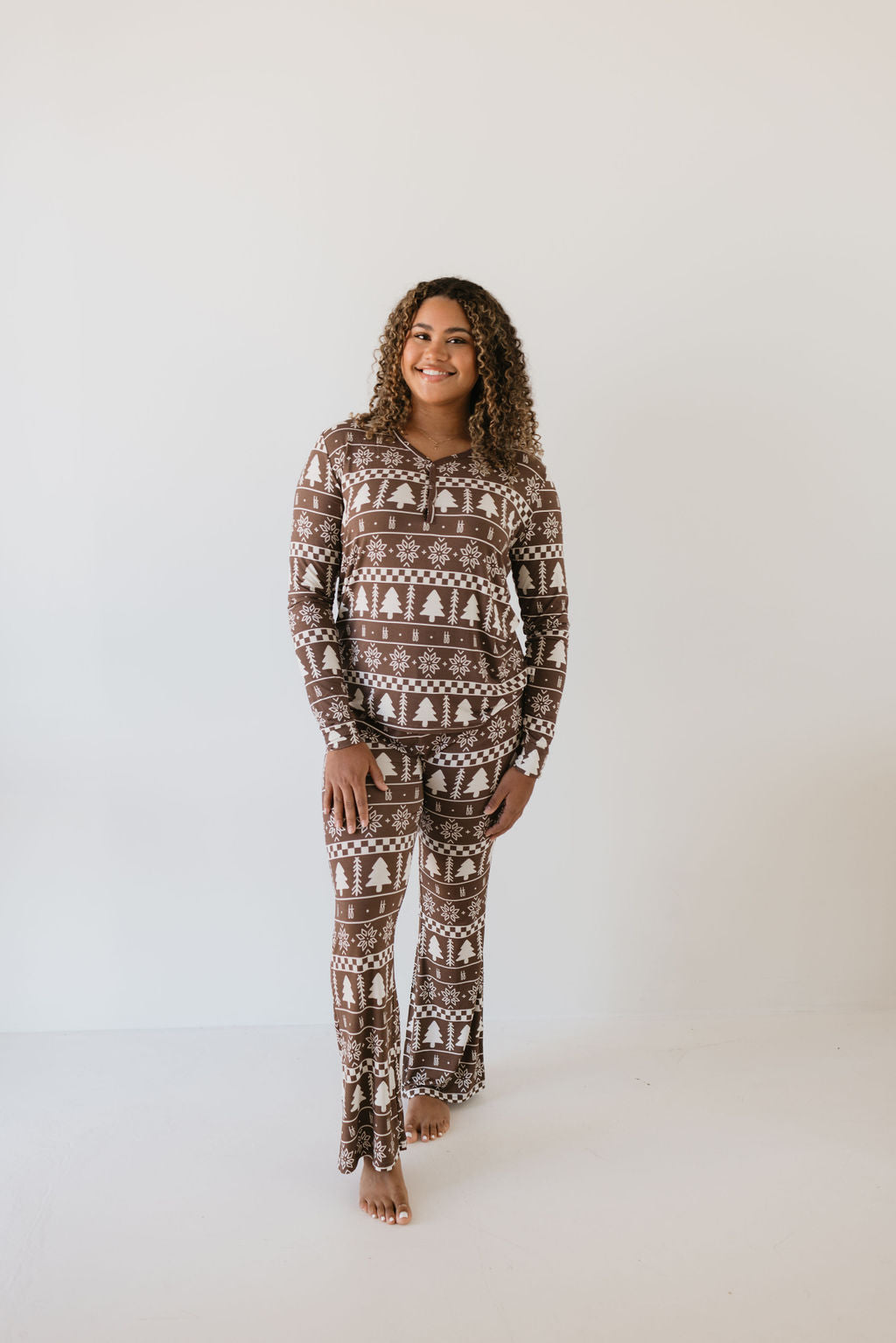 Against a simple white backdrop, someone is shown barefoot and smiling while wearing women's bamboo pajamas by forever french baby. The cozy spirit of the season is captured perfectly with their "Forever Fair Isle" holiday pattern featuring trees and snowflakes.