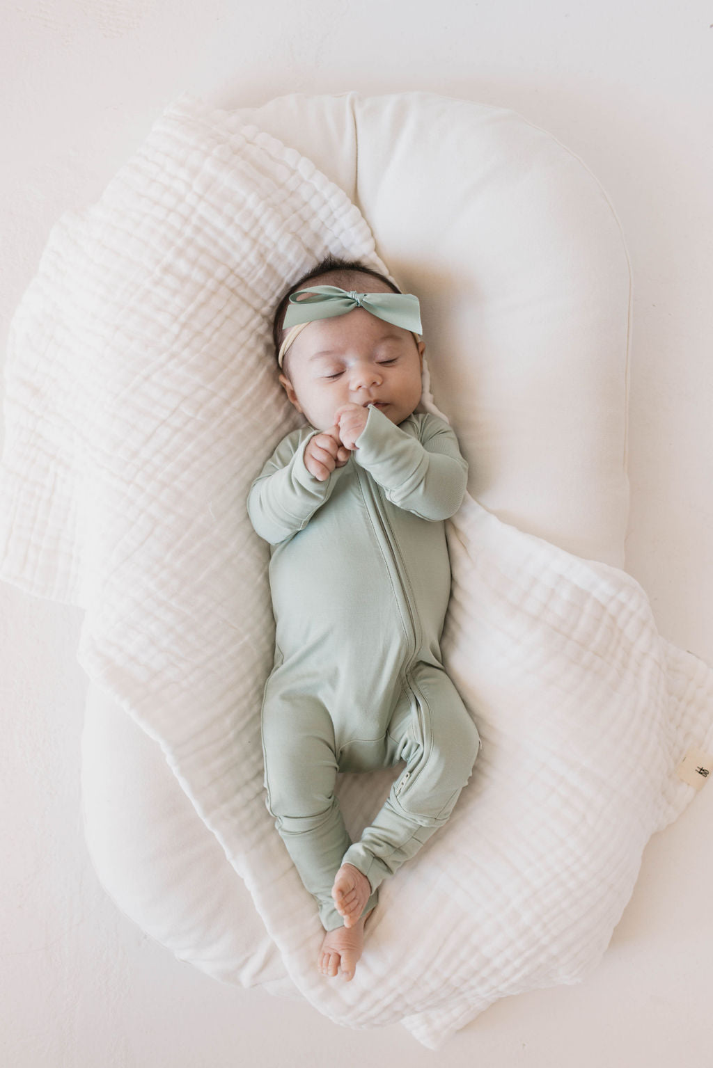 A sleeping baby wearing Forever French Baby's Bamboo Zip Pajamas in Pistachio, complete with a matching bow headband, is lying on a cushioned white blanket. The hypo-allergenic pajamas ensure the baby's comfort and, with hands clasped together, they appear peaceful and content on the softly textured blanket that cradles them gently.