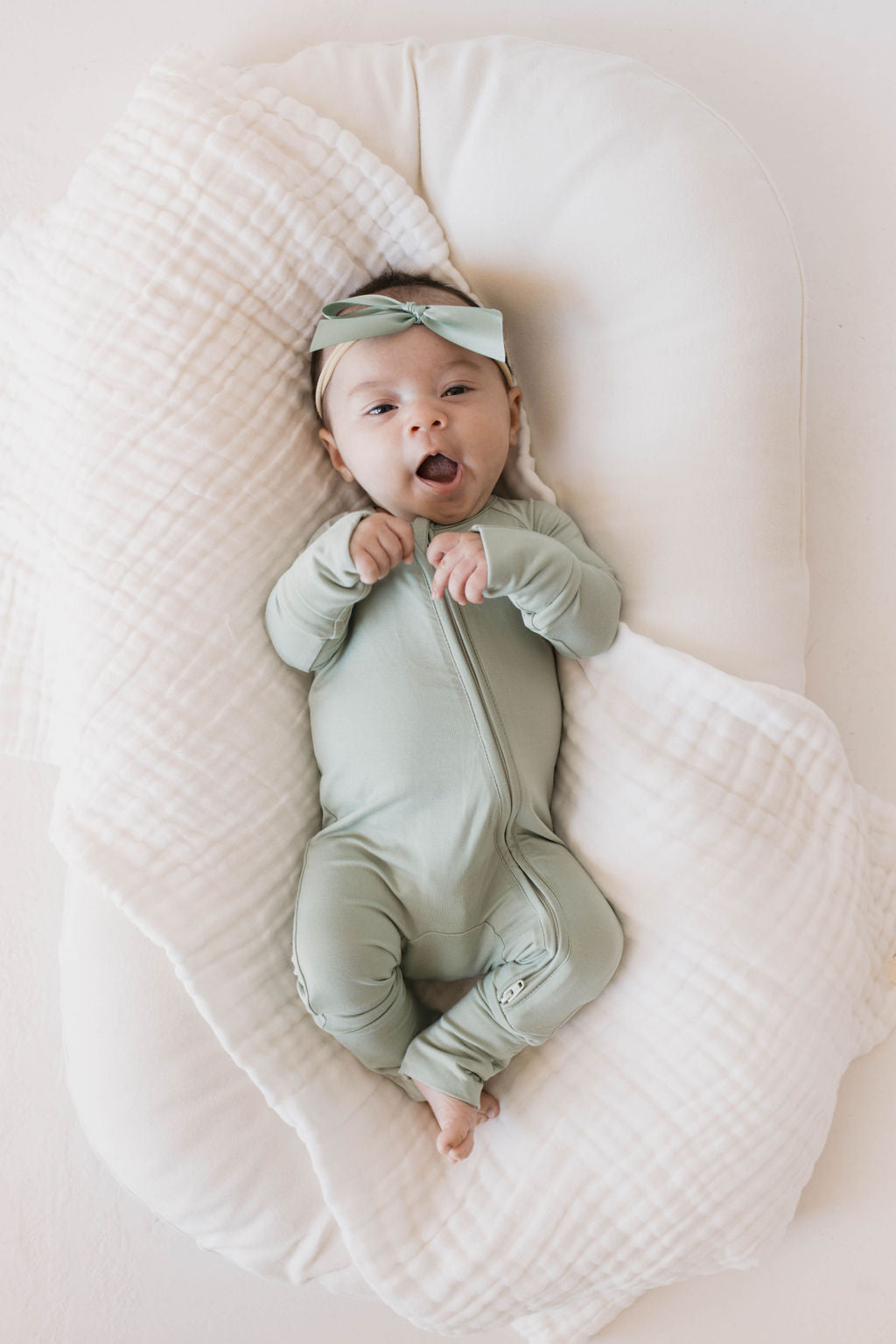 A baby wearing forever french baby's Bamboo Zip Pajamas in Pistachio, complete with a matching headband, lies on a white cushioned surface with a soft, textured white blanket. The baby has an open mouth and appears to be smiling or cooing.