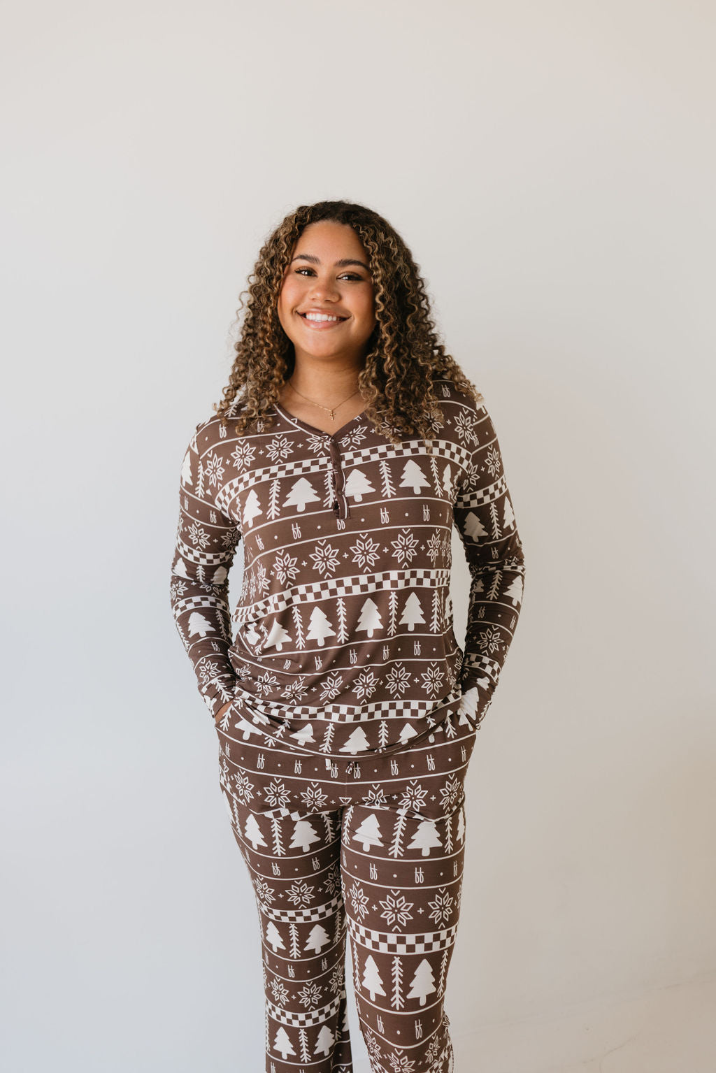 A person smiling in the Women's Bamboo Pajamas from Forever French Baby, named "Forever Fair Isle," stands against a plain white background. The brown and white pajama set features winter-themed patterns with snowflakes and pine trees, designed to ensure comfort and style for those with sensitive skin.