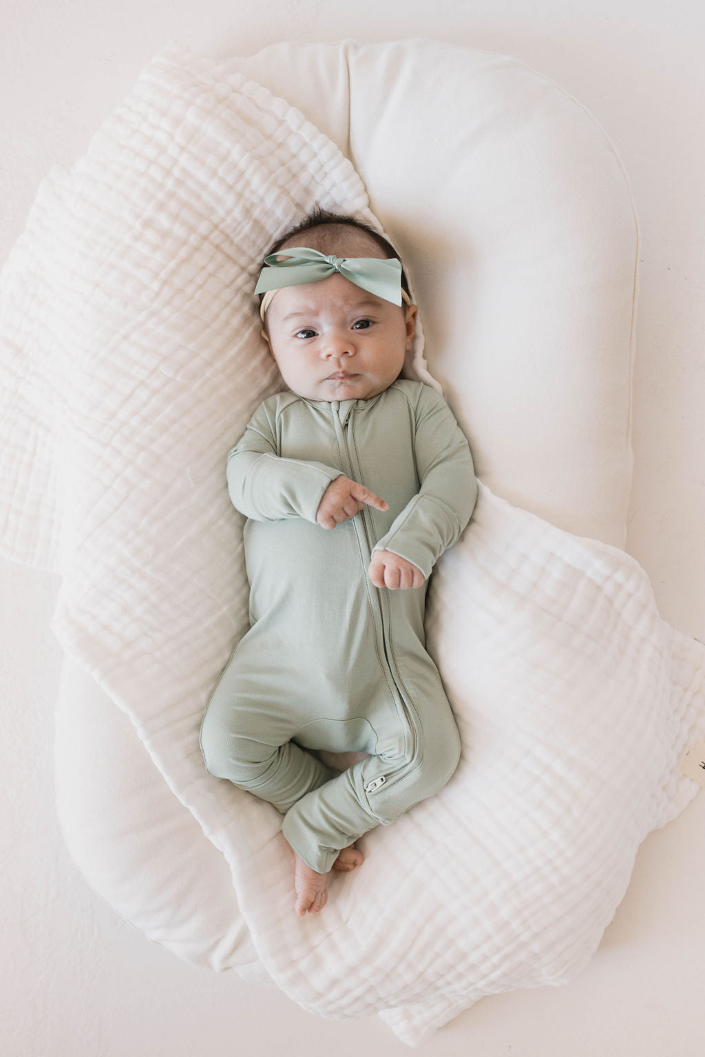 A baby wearing pistachio Bamboo Zip Pajamas from forever french baby rests on a white cushioned surface, adorned with a matching headband. The infant gazes directly at the camera, both arms resting on the hypoallergenic, textured blanket.