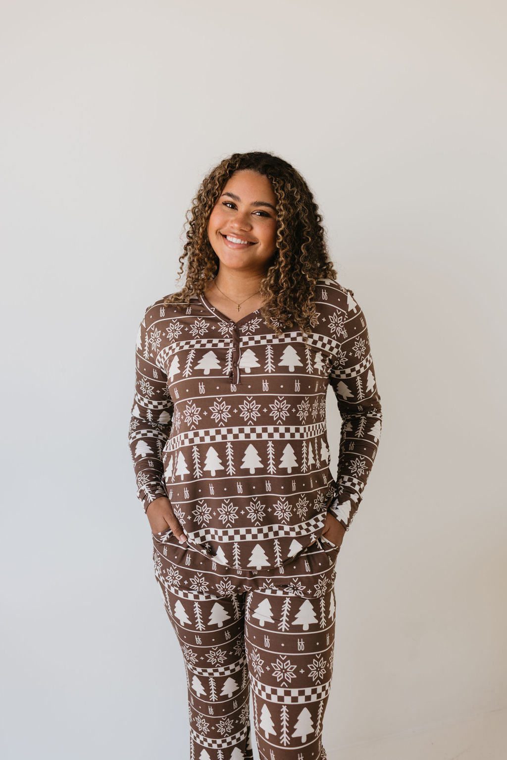 A person, wearing Forever Fair Isle women's pajamas from forever french baby, smiles against a light background. These stylish bamboo pajamas feature white winter-themed patterns like snowflakes and trees, with the individual comfortably placing hands in pockets. The matching baby and toddler pajamas nearby ensure hypo-allergenic comfort for everyone.