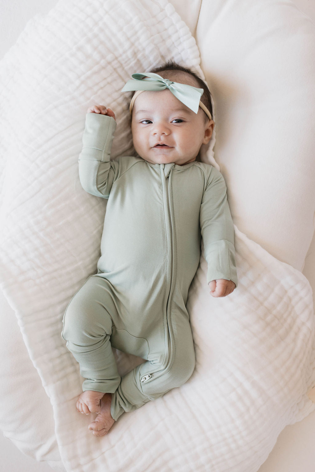 A baby wearing bamboo zip pajamas in pistachio from Forever French Baby and a matching headband with a bow is lying on a soft, white quilted surface. The hypo-allergenic clothing ensures comfort as the baby’s arm is slightly raised, looking into the camera with a subtle smile.