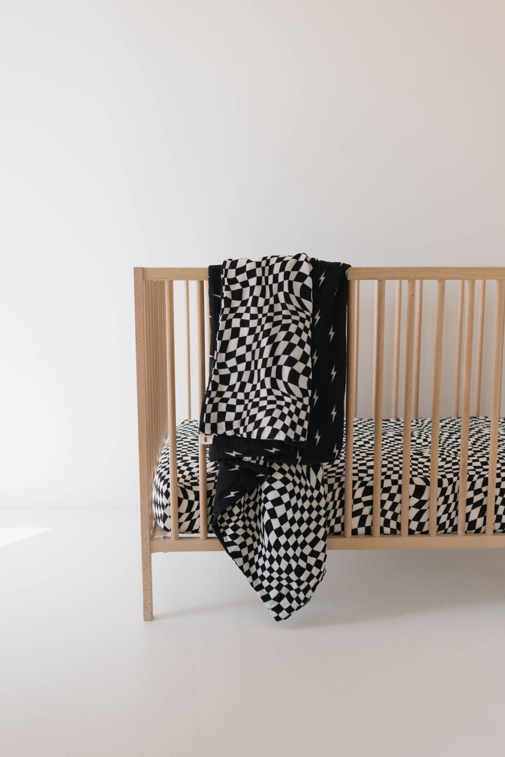 A minimalist nursery setup featuring a wooden crib adorned with the Muslin Quilt by forever french baby, showcasing Midnight Lightning Bolt and Black + White Wavy Checkerboard patterns. Matching muslin cotton blankets are draped over the side. The room has a clean, white background, enhancing the modern aesthetic.