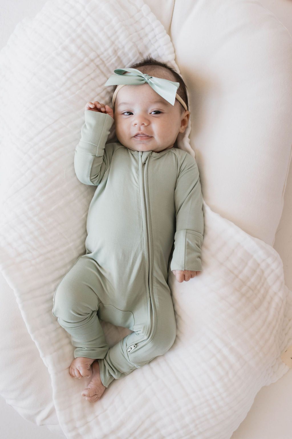 A baby dressed in Forever French Baby’s Bamboo Zip Pajamas in Pistachio, complete with a matching headband and bow, lies on a white quilted blanket. The baby gazes upwards, one hand raised near their face with their bare feet resting comfortably. The entire scene exudes a soft and serene ambiance, highlighting the luxurious and hypo-allergenic qualities of Forever French Baby's premium clothing line.