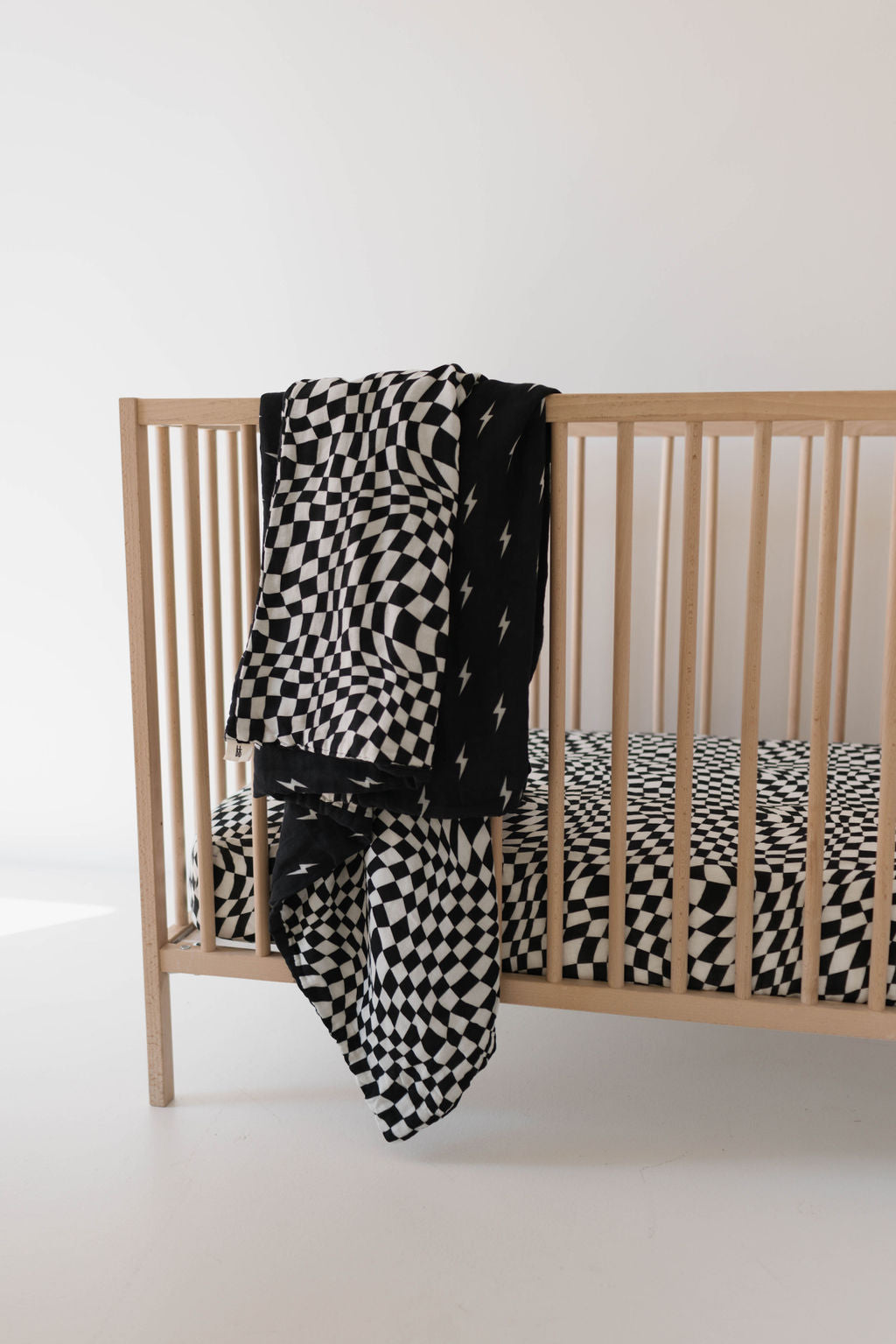 A wooden baby crib featuring the Muslin Quilt | Midnight Lightning Bolt & Black + White Wavy Checkerboard from forever french baby draped over its side. The crib mattress, dressed in matching nursery bedding, showcases the same black and white geometric design. The room is minimalistic and well-lit.