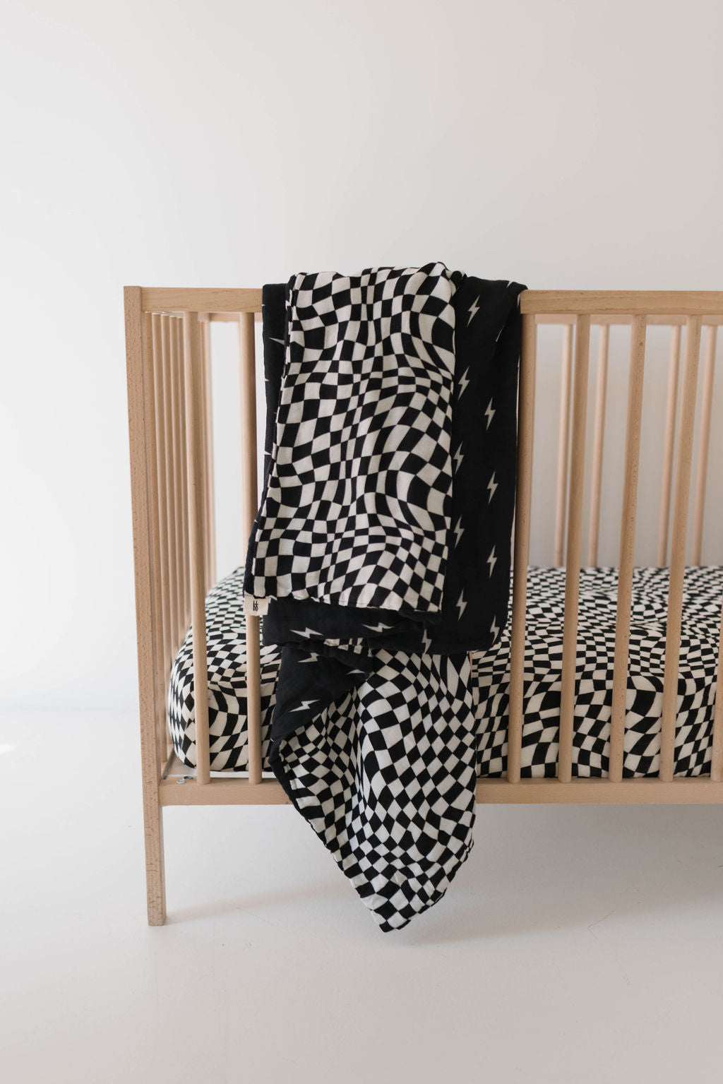 A wooden baby crib, adorned with black and white wavy checkerboard nursery bedding from the forever french baby collection, features a delightful Muslin Quilt in Midnight Lightning Bolt & Black + White Wavy Checkerboard draped over its side. The minimalistic design of the room is further enhanced by the plain white wall in the background.