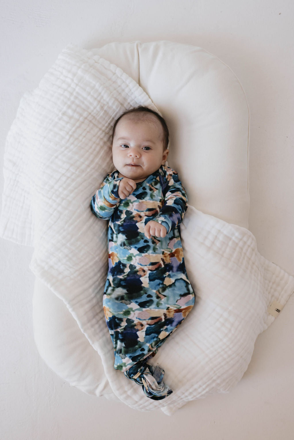 A baby dressed in the vibrant Charli Print Bamboo Knotted Gown from Forever French Baby lies on a white, cushioned pad with a textured white blanket partially covering it. The baby has a curious expression, their left hand near their mouth. Supporting autism awareness, the background is a smooth, light-colored surface.