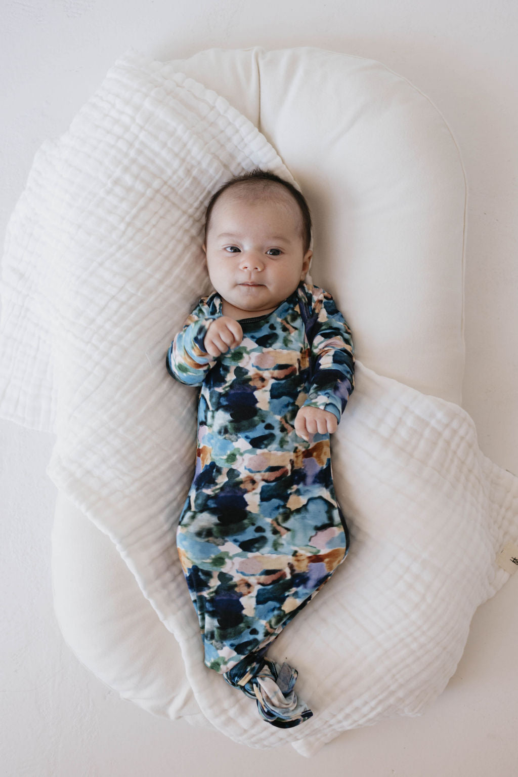 A baby wearing the forever french baby Bamboo Knotted Gown in Charli Print lies on a white cushioned surface with a soft, white blanket partially covering them. The baby gazes at the camera with a calm expression.