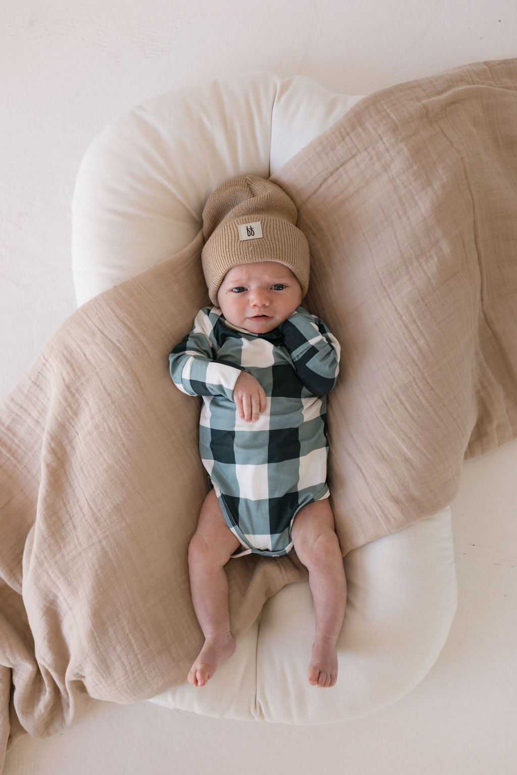 A baby in a Long Sleeve Bamboo Snapsuit in Evergreen Gingham from forever french baby and a beige knit hat rests on a soft white cushion, swaddled in a light brown blanket made of hypo-allergenic bamboo fabric. They gaze upwards with a peaceful expression.