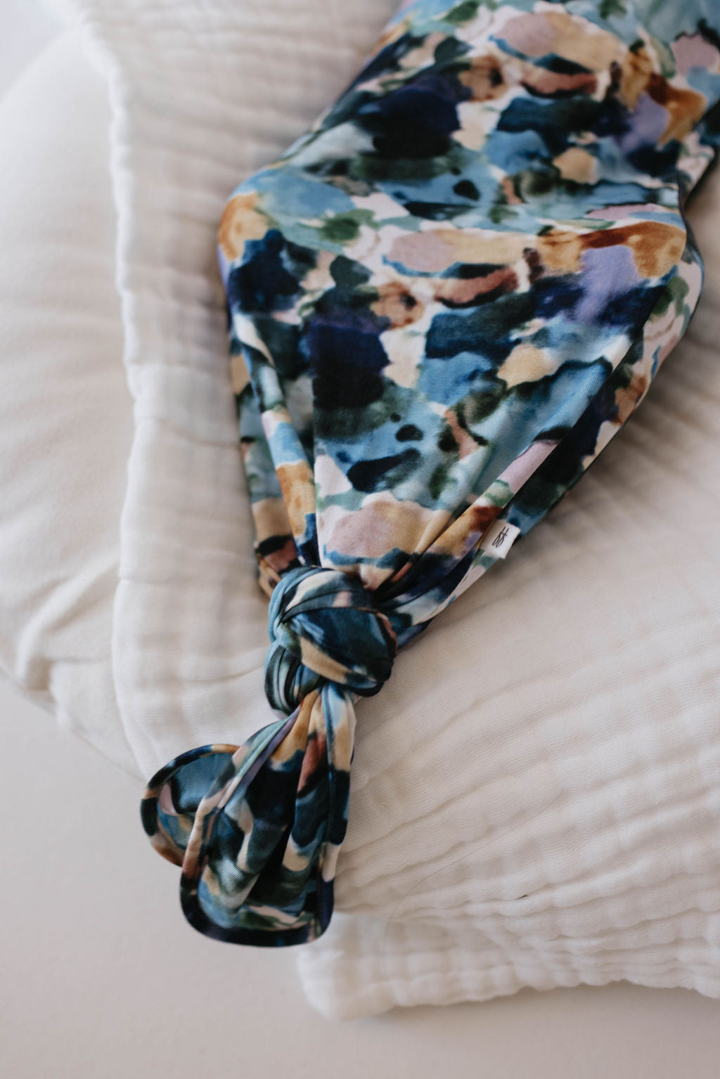 A soft and colorful Bamboo Knotted Gown in Charli Print, featuring a blue, green, and brown abstract pattern from forever french baby, is loosely tied in a knot and placed on a quilted white fabric background. The design evokes the artistic touch of an autistic daughter’s creations.