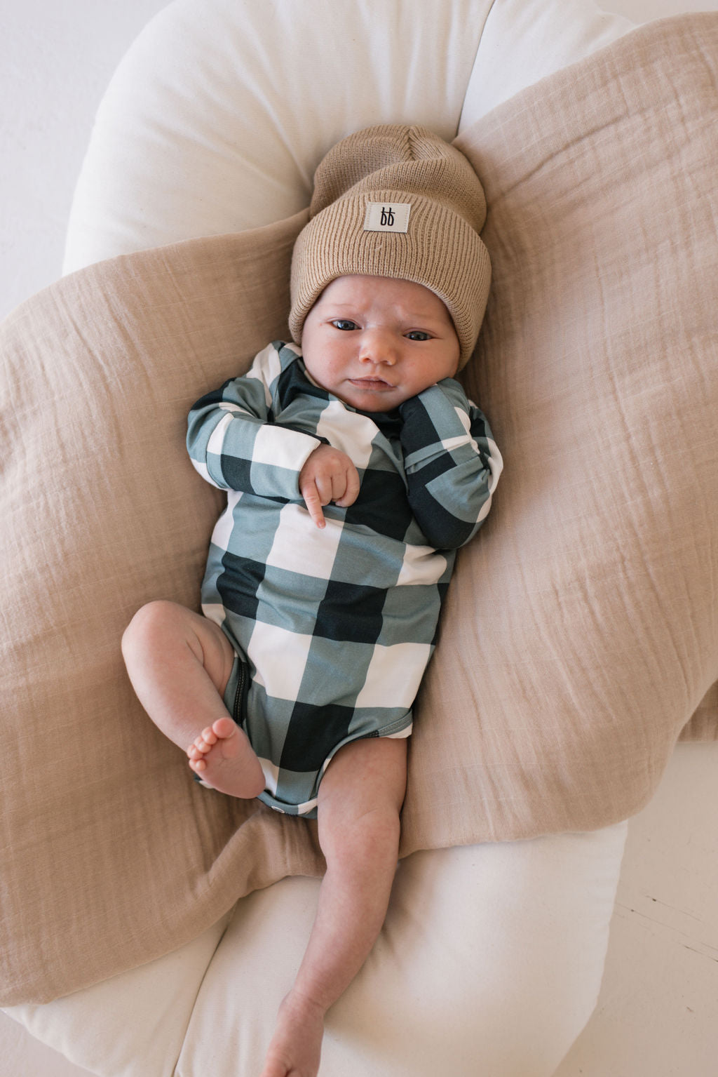 A baby wearing the evergreen gingham long sleeve bamboo snapsuit by Forever French Baby, paired with a beige knit hat, lies on a cozy, cushioned surface. Nestled between soft, neutral-colored blankets, the baby has a gentle and relaxed expression.