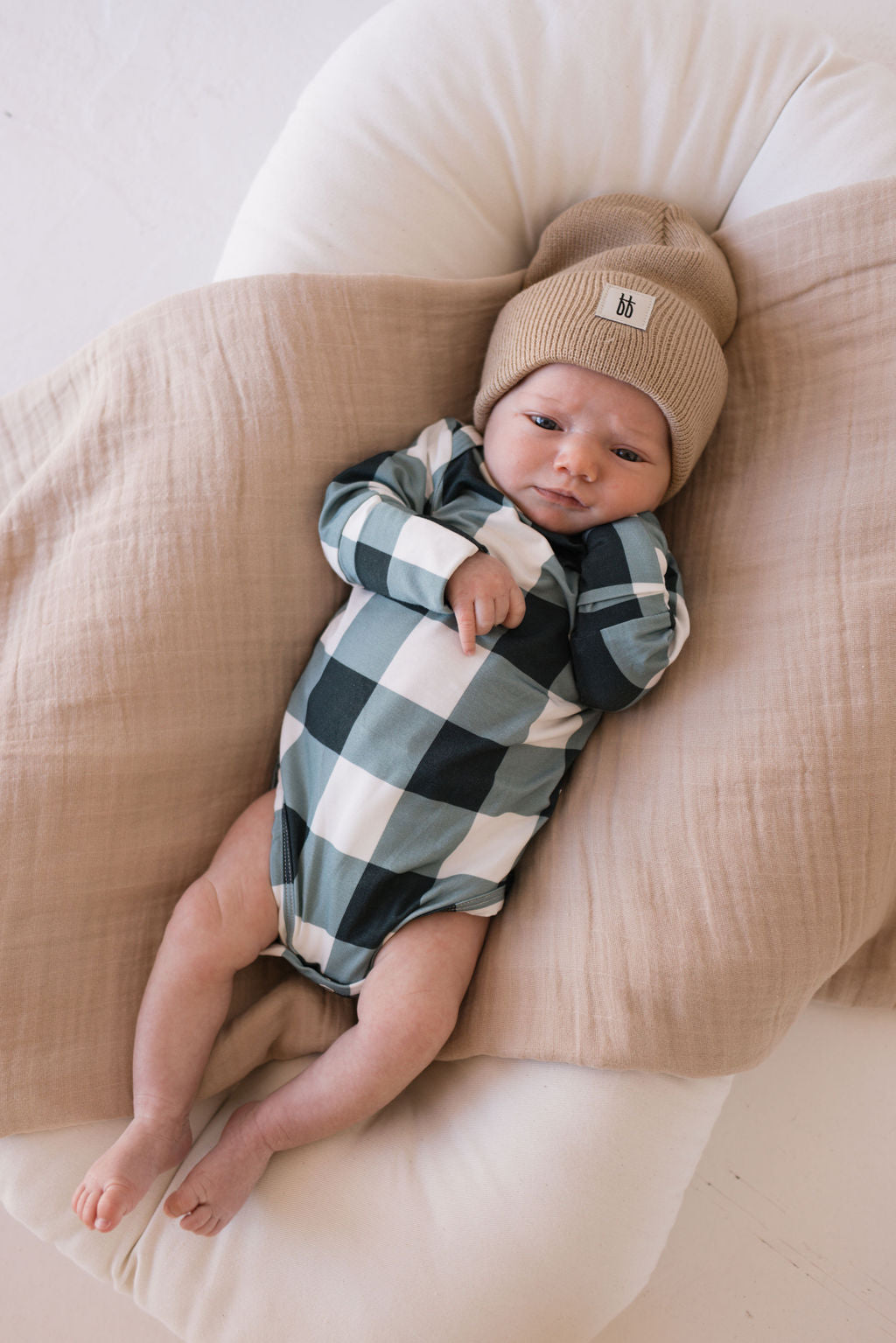 A baby lies peacefully on a cushioned surface, dressed in the Long Sleeve Bamboo Snapsuit in Evergreen Gingham by forever french baby and a beige knit beanie. Wrapped in a cozy beige blanket, the little one gazes at the camera with a calm expression.