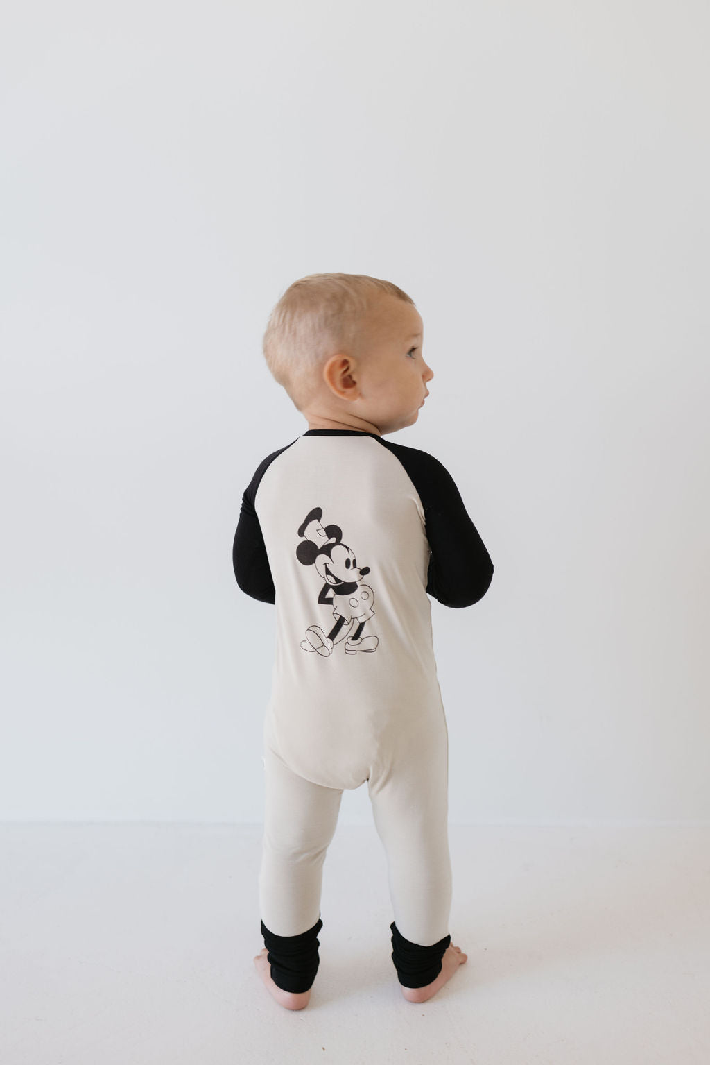 A toddler stands facing away, dressed in forever french baby's Bamboo Zip Pajamas | Steamboat Willie. The beige onesie with black sleeves and legs is made from breathable fabric and showcases a large cartoon mouse character on the back, set against a plain white background.