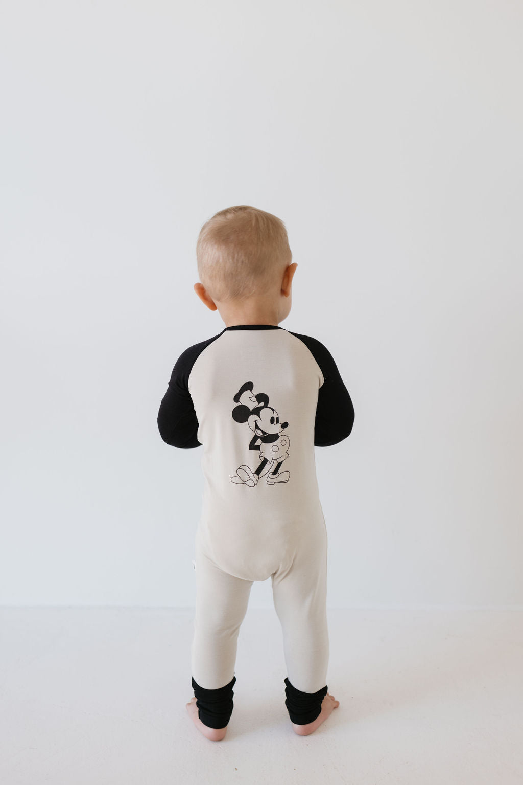 A baby wearing the forever french baby Bamboo Zip Pajamas in cream and black, made from breathable fabric, stands with their back to the camera. The pajamas feature a Steamboat Willie cartoon character graphic on the back. Set against a plain white background, they showcase the comfort and style of hypo-allergenic clothing.