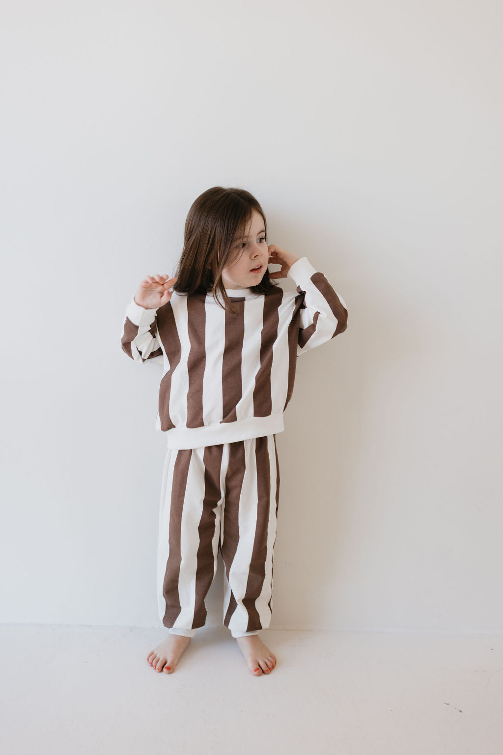 A young child with long dark hair stands barefoot against a light background, wearing a cozy Sweat Set from forever french baby in the Coffee Shoppe style, featuring vertical brown and white stripes.