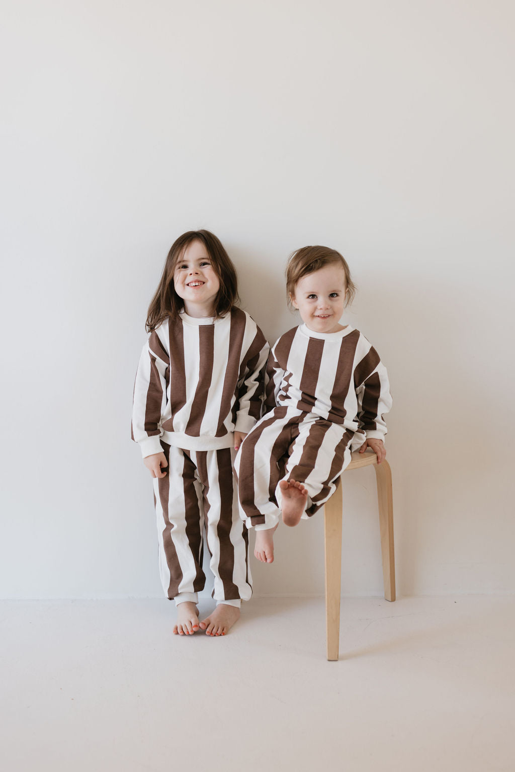 Two children dressed in the "Sweat Set | Coffee Shoppe" by forever french baby, featuring brown and white stripes. One child is seated on a stool while the other stands nearby, both against a plain white wall. Barefoot and smiling, they appear perfectly cozy in their cooler weather outfits.