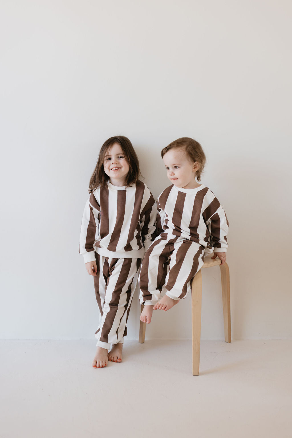 Two young children wearing matching Coffee Shoppe sweat sets by forever french baby. One child is seated on a wooden stool while the other stands beside, both smiling in a white-themed room, perfectly dressed for cooler weather.