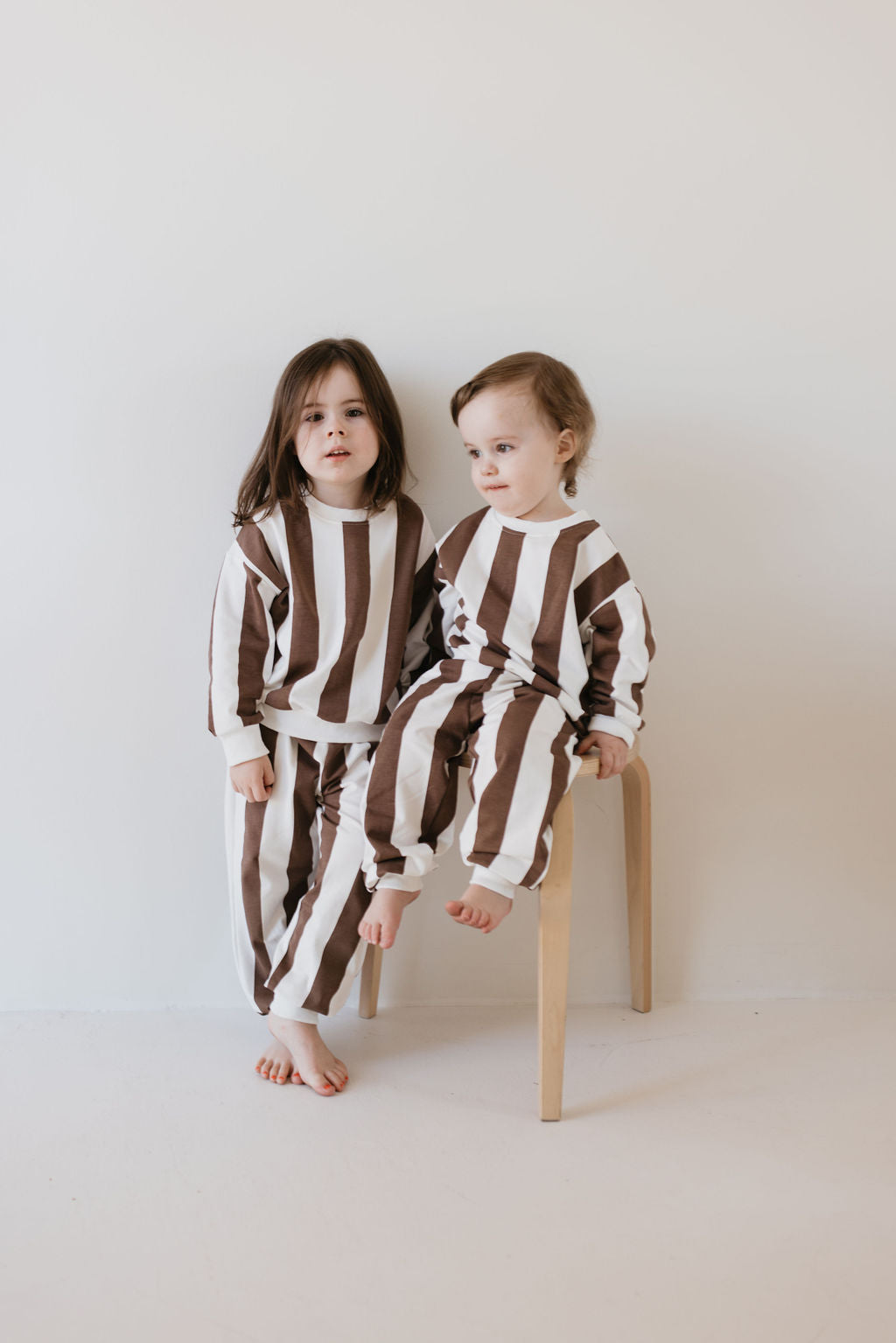 Dressed in the stylish "Sweat Set | Coffee Shoppe" by forever french baby, two young children create a charming scene. One sits on a wooden chair while the other stands beside them, both barefoot against a plain, light-colored background.