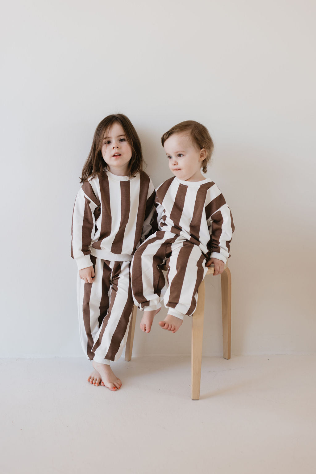 Two young children are wearing matching "Sweat Set | Coffee Shoppe" outfits by forever french baby. One stands while the other sits on a wooden stool, making them look adorable for cooler weather outings to the local coffee shoppe, all set against a plain white background.