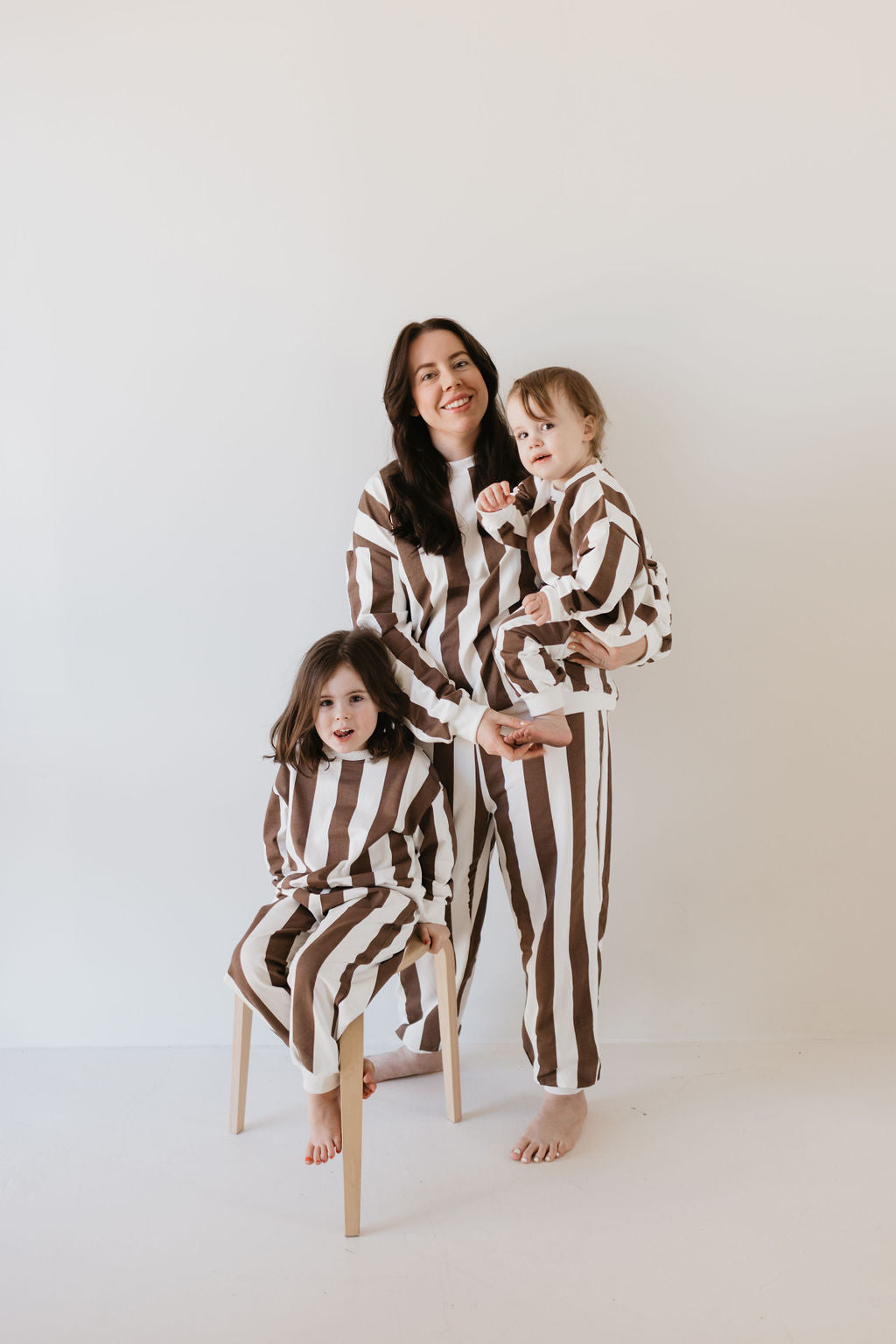 A woman and two children wearing the Sweat Set | Coffee Shoppe by forever french baby gather together, perfectly capturing the cozy essence of cooler weather attire. One child sits on a wooden stool, while the other is affectionately held by the woman in their minimalist, light-colored room.