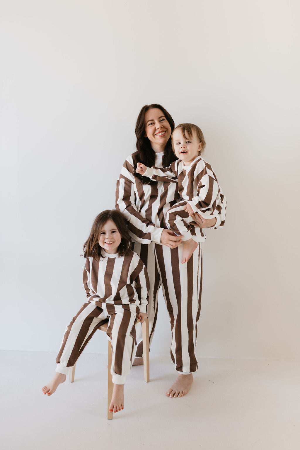 A woman and two children are dressed in matching "Sweat Set | Coffee Shoppe" outfits by forever french baby, ideal for cooler weather. The woman holds a toddler as the other child sits on a stool, all smiling against a plain light background.