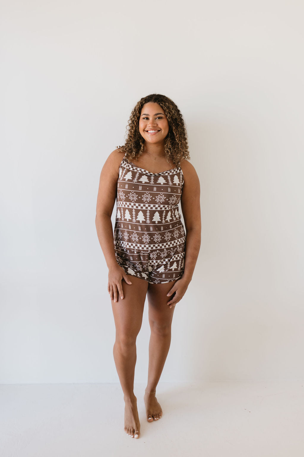 A person with curly hair cascading around their shoulders stands barefoot against a plain background, smiling. They are dressed in the Cami Women's Bamboo Set from forever french baby, featuring a brown tank top and shorts adorned with a white winter-themed pattern of trees and snowflakes, crafted from breathable bamboo fabric.