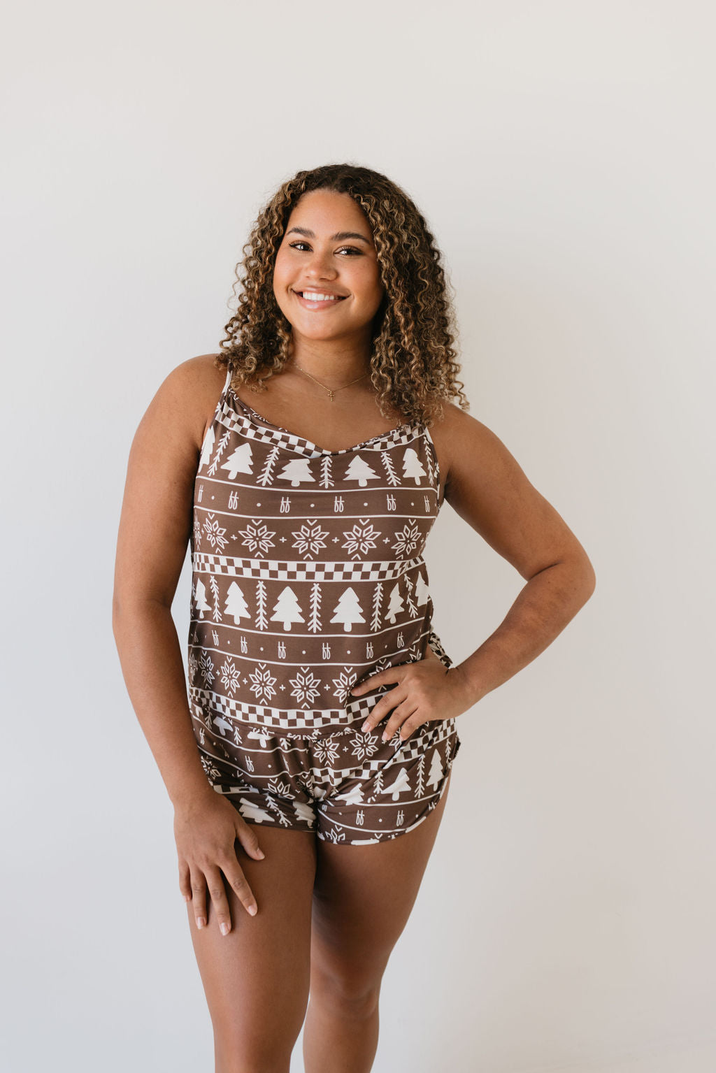 A person with curly hair is smiling while wearing the Cami Women's Bamboo Set in Forever Fair Isle by forever french baby. The brown pajamas, made from breathable bamboo fabric, feature white holiday-themed patterns like trees and snowflakes against a plain light background.