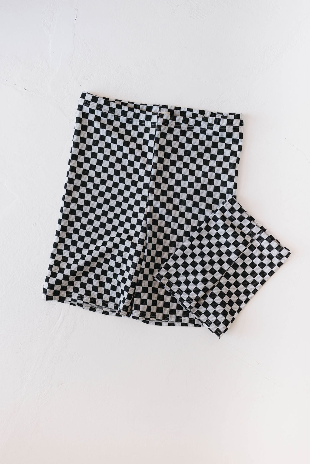 A pair of Women's Biker Shorts in black and white ribbed checker from forever french baby is neatly folded beside a matching checkered face mask. Both small items are placed on a plain, white surface, and they are machine wash friendly for easy care.