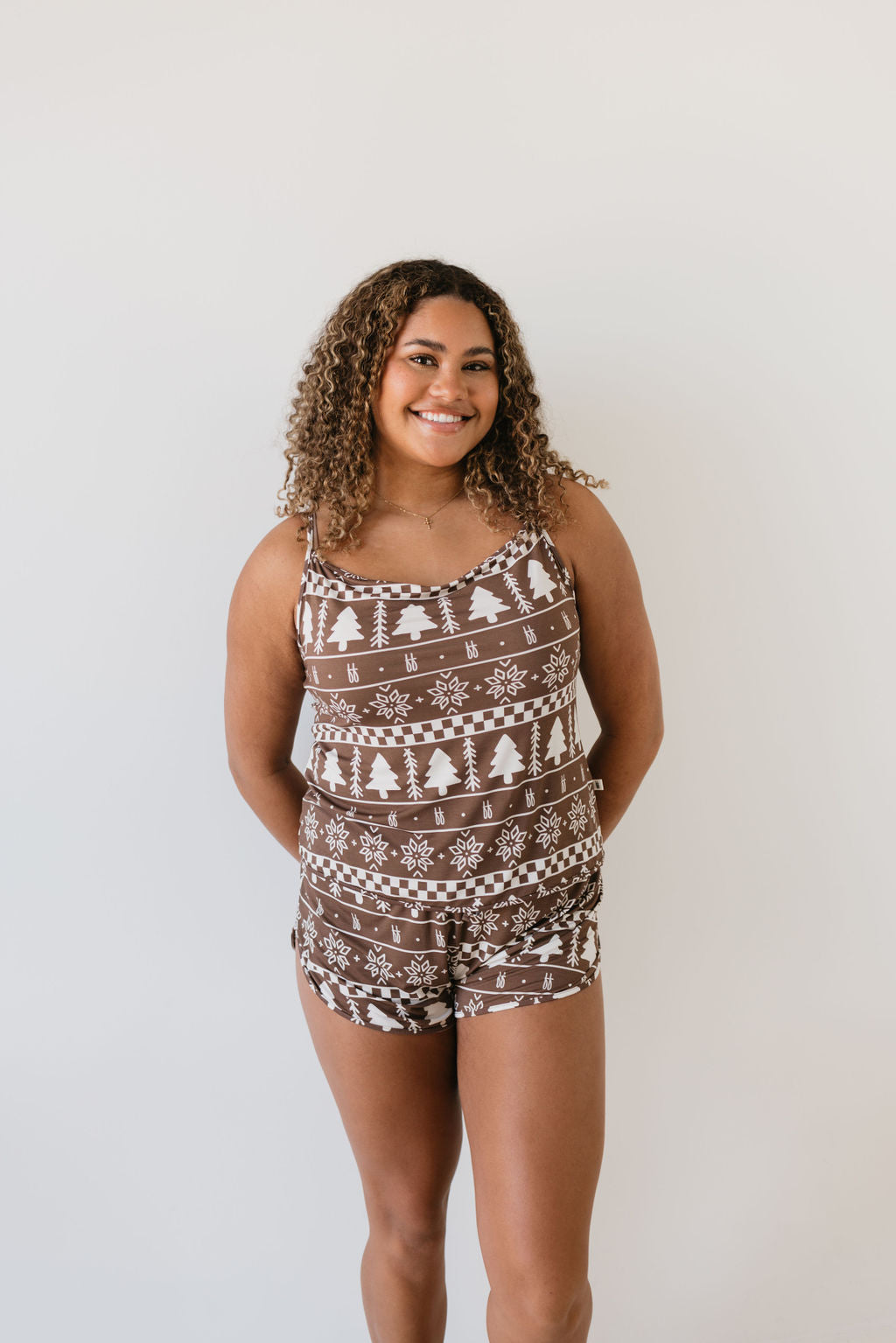 A woman models the Cami Women's Bamboo Set from Forever French Baby, featuring a brown tank top and shorts adorned with white festive patterns, while smiling against a simple backdrop.