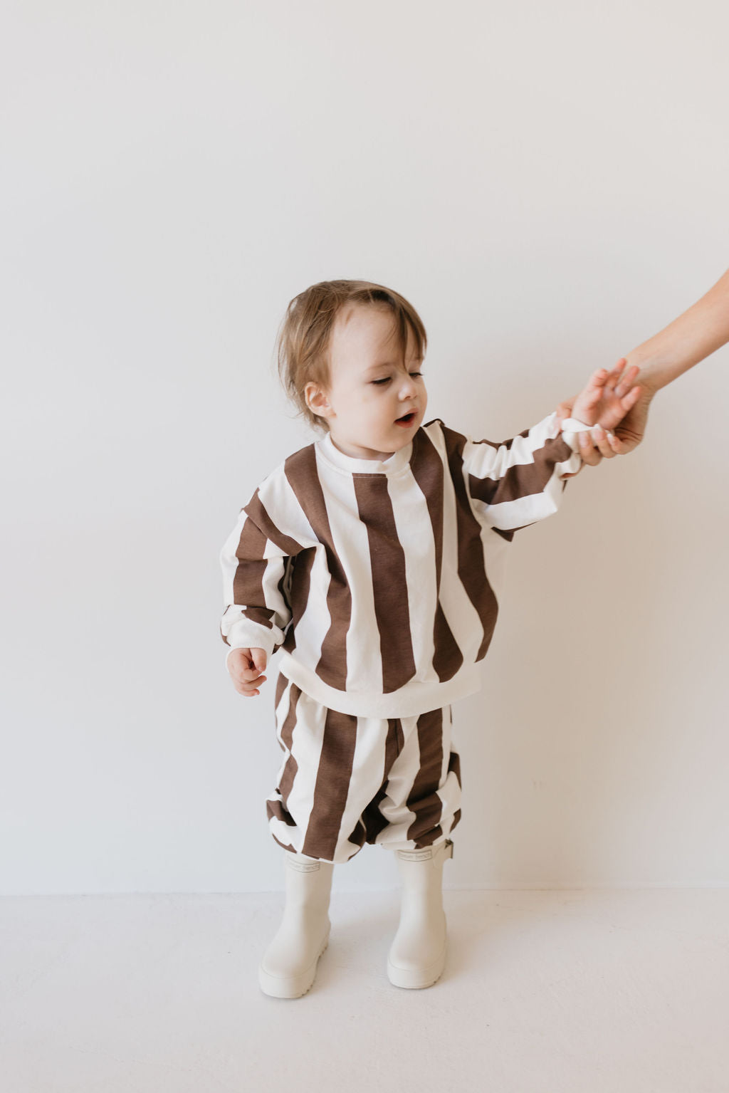 A toddler in a Sweat Set | Coffee Shoppe by forever french baby, featuring a white and brown striped outfit and white boots for cooler weather, holds an adult's hand against a plain, light-colored background.