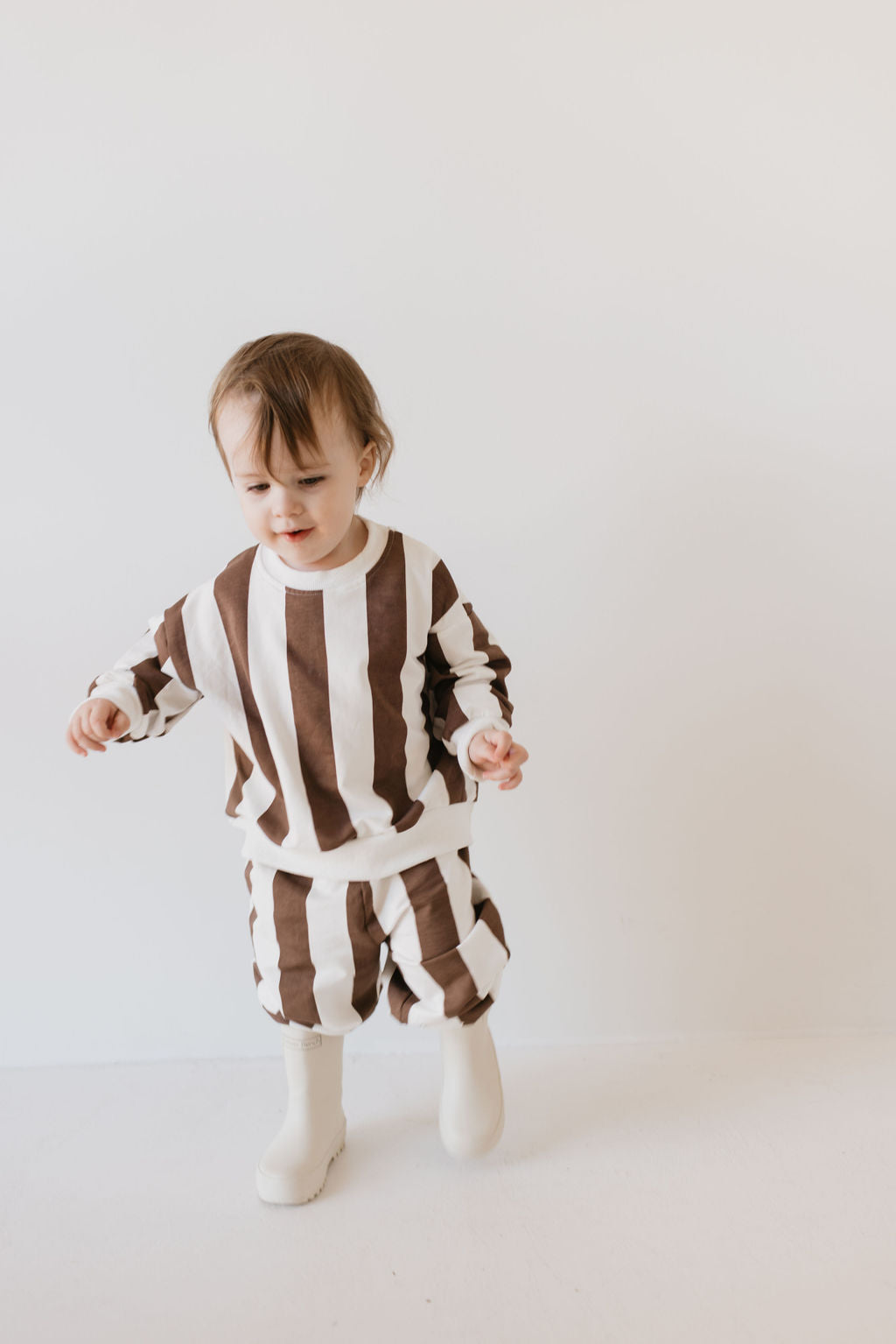 A toddler dressed in a Sweat Set from the Coffee Shoppe collection by forever french baby stands against a simple light background, adorably wearing white boots. The child looks as if they are taking a cautious step forward, perfectly suited for cooler weather adventures.