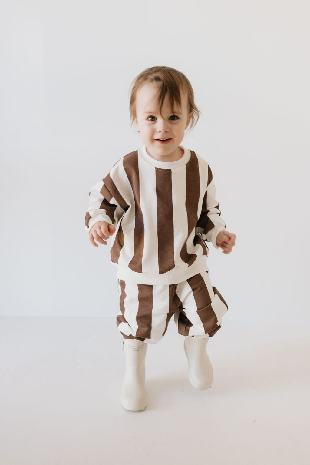 A toddler is beaming in a cozy "Sweat Set | Coffee Shoppe" by forever french baby, featuring large vertical brown and white stripes. Against a plain white backdrop, the little one sports short hair while wearing the long-sleeve top and pants paired with white boots—making it perfect for cooler weather.