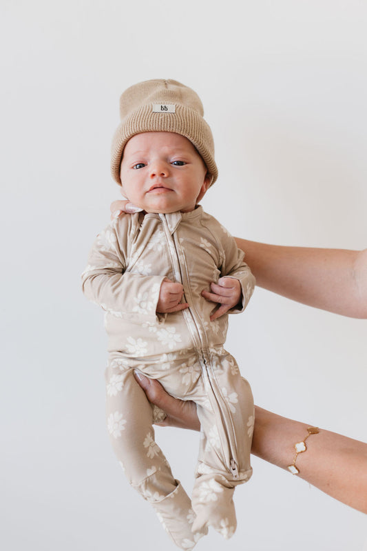 A baby dressed in the "Bamboo Zip Pajamas | Lazy Daisy" from Forever French Baby, featuring a brown base adorned with white floral patterns and accompanied by a matching beanie, is gently held up by an adult's hands against a plain, light background.