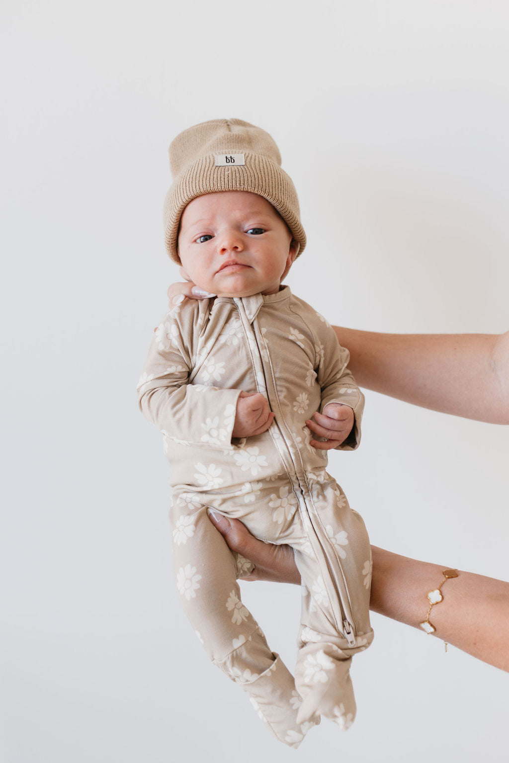 A baby wearing the Bamboo Zip Pajamas in the Lazy Daisy pattern, accompanied by a matching beanie from forever french baby, is held against a plain white background. The baby's calm expression meets the camera, exuding comfort and serenity.