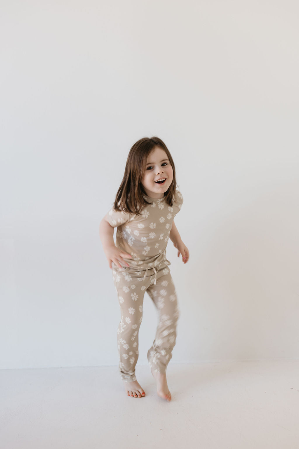 A young girl with long brown hair is smiling while wearing the Toddler Short Sleeve Bamboo Lounge Set in the Lazy Daisy pattern by forever french baby. The beige outfit, made from breathable bamboo fabric with white floral designs, adds to her playful demeanor as she poses joyfully against a plain white background.