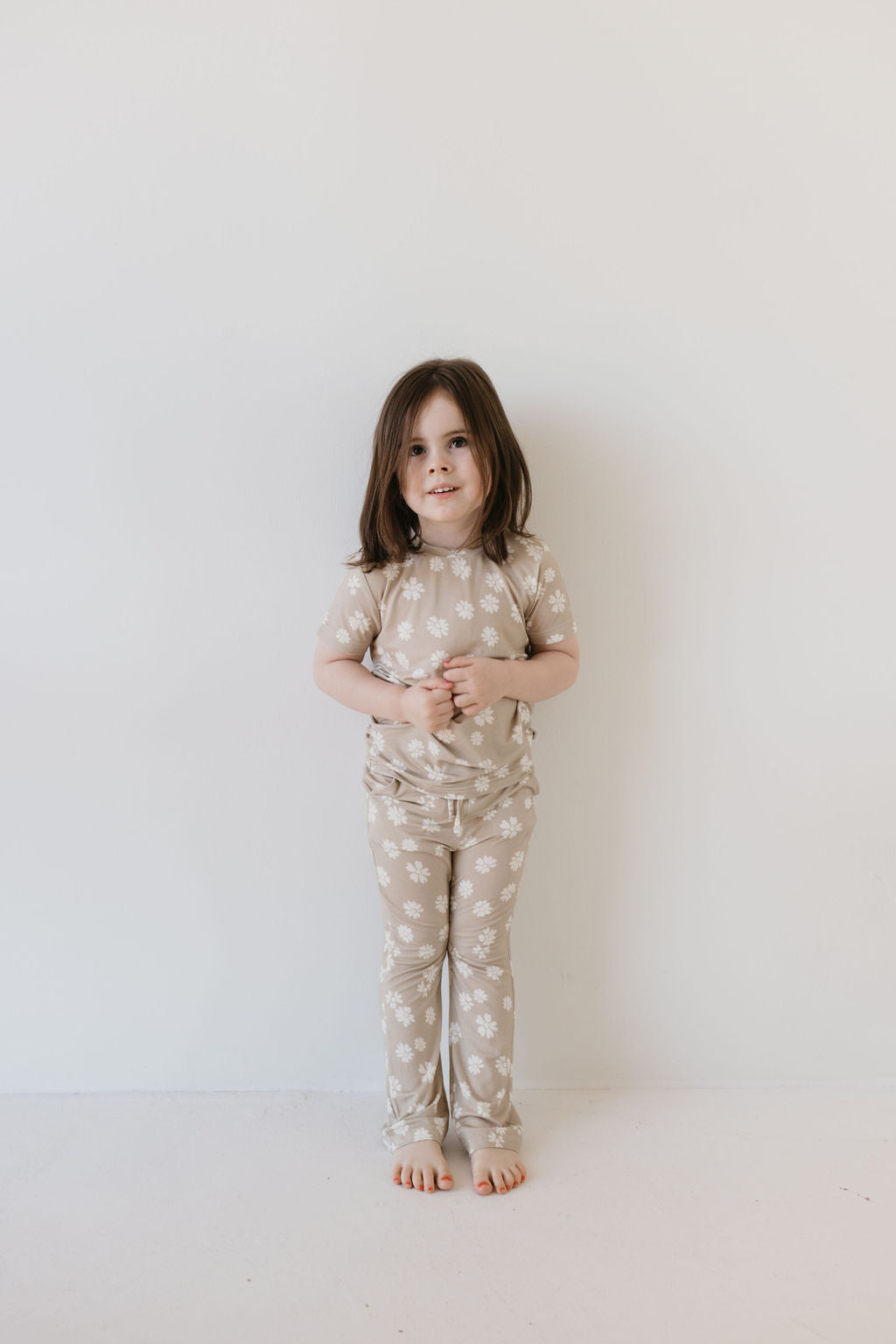 A young child with long hair stands barefoot against a plain light-colored wall, wearing the Toddler Short Sleeve Bamboo Lounge Set in the Lazy Daisy pattern from forever french baby. The outfit is made from breathable, hypo-allergenic bamboo and features a charming floral design. The child gazes at the camera with hands clasped.