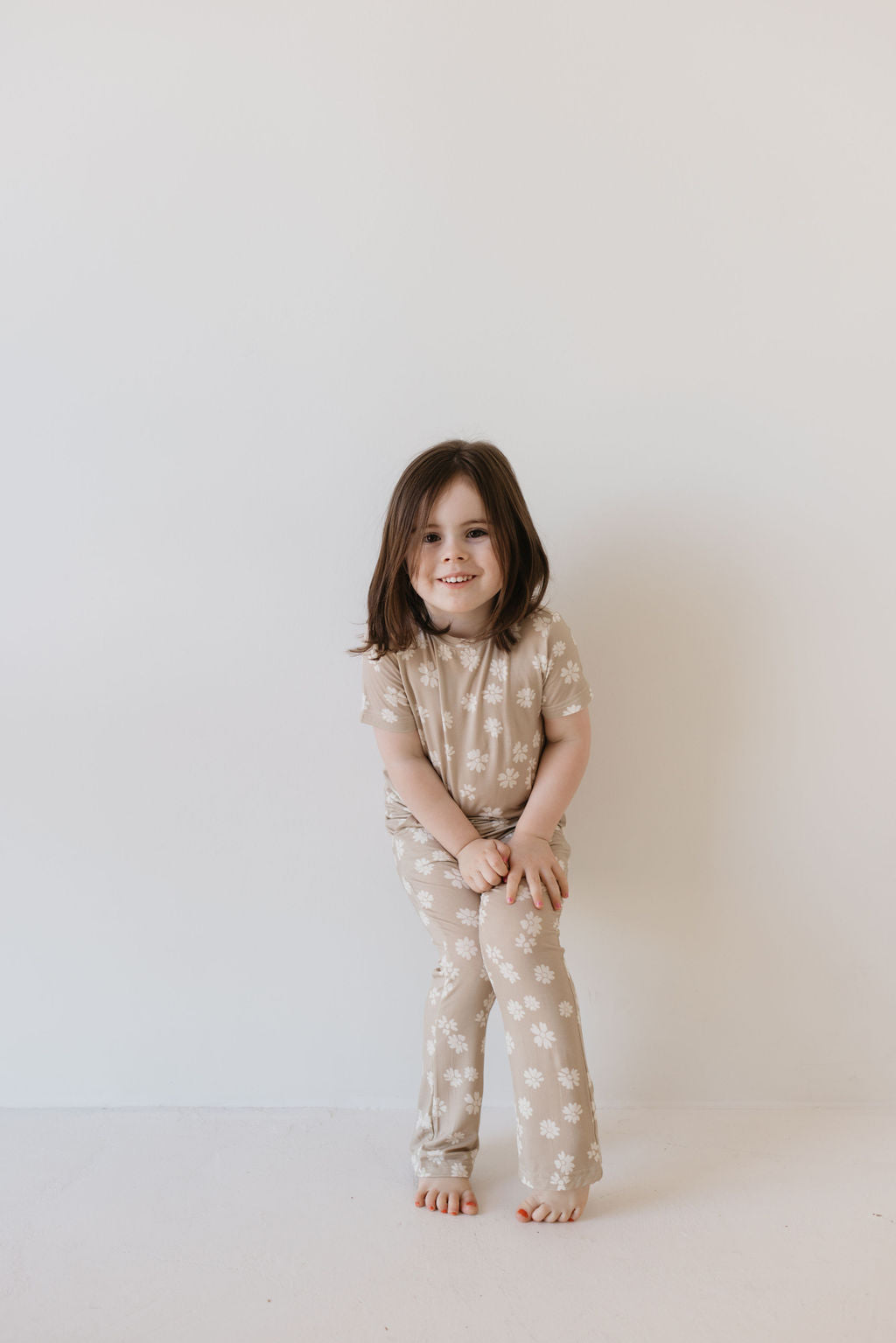 A young child with long brown hair stands against a plain white wall, wearing the Toddler Short Sleeve Bamboo Lounge Set in the Lazy Daisy design by forever french baby. The breathable beige pajamas feature a charming white floral pattern, and the child smiles while leaning slightly forward, legs crossed and hands resting on their knees.