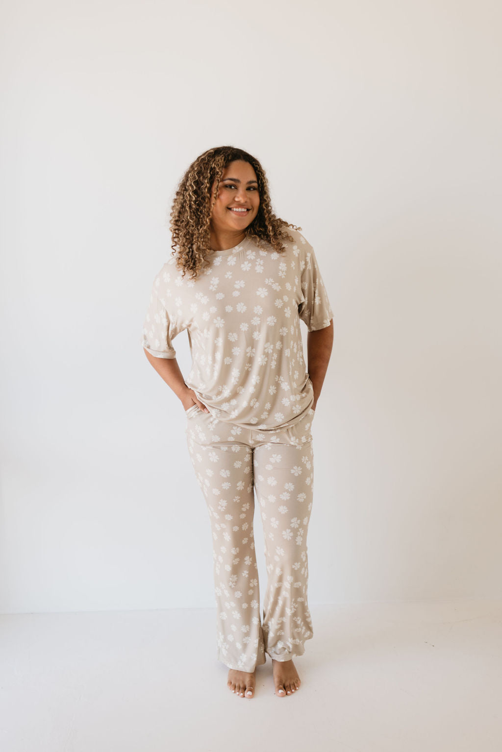 A person with curly hair is smiling at the camera while standing barefoot against a plain white background. They are wearing the oversized Lazy Daisy short sleeve women's bamboo pajamas from forever french baby, featuring a white floral pattern. The hypoallergenic fabric provides both comfort and style.