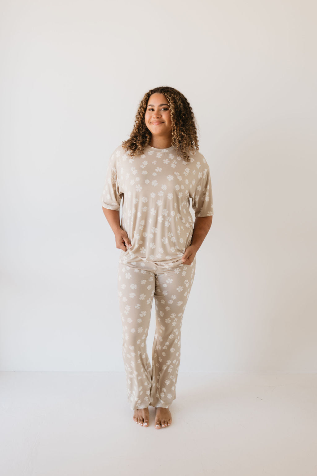 A person with curly hair stands barefoot against a white background, smiling gently in a relaxed posture while wearing the Lazy Daisy Short Sleeve Women's Bamboo Pajamas by forever french baby. The oversized pajama set features a subtle floral pattern in beige and is hypo-allergenic.