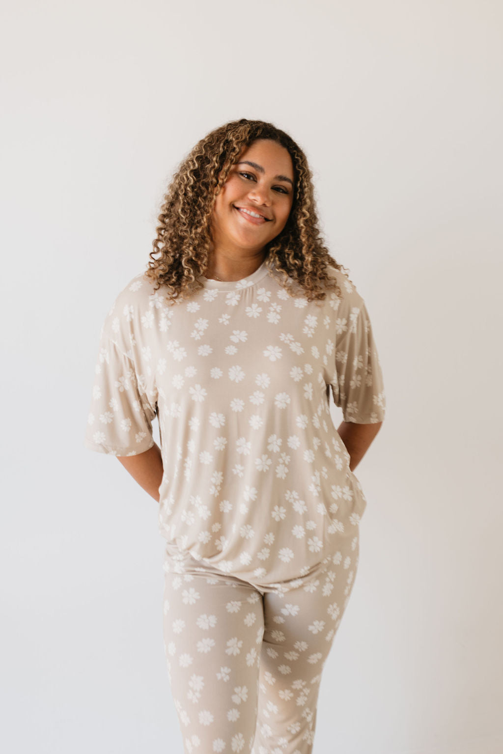 A person with curly hair smiles while standing against a plain background. They are wearing the light-colored Short Sleeve Women's Bamboo Pajamas from the Lazy Daisy collection by forever french baby, which features a white floral pattern and is known for its hypo-allergenic properties.