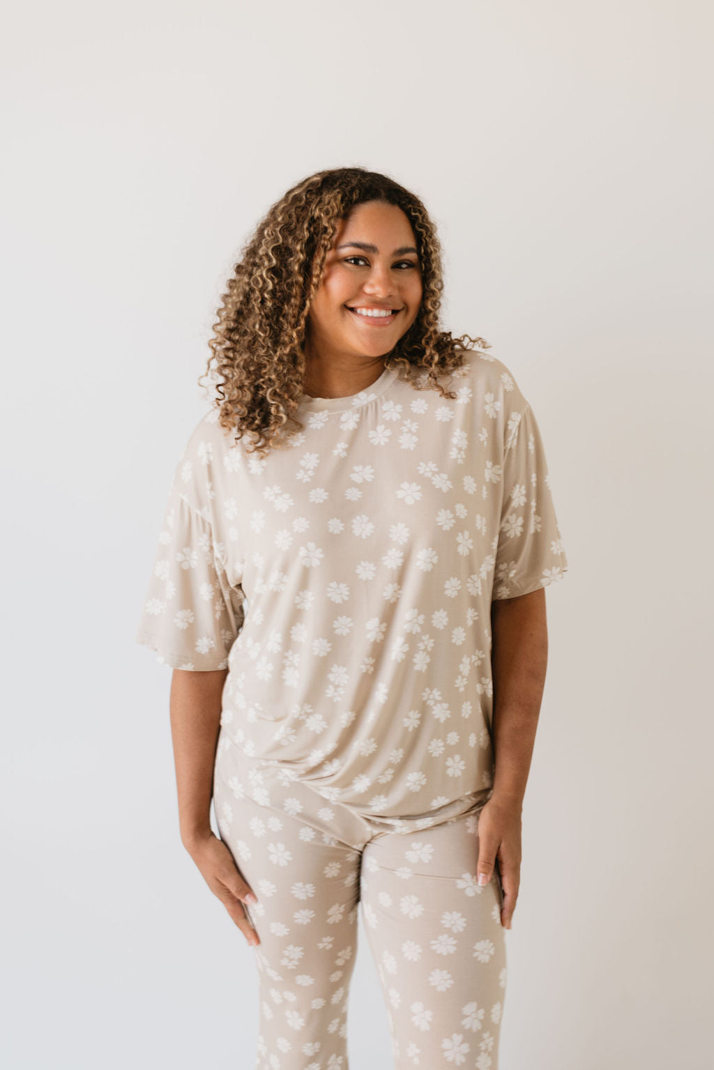 A smiling woman with curly hair is wearing the Short Sleeve Women's Bamboo Bamboo Pajamas in the Lazy Daisy design by Forever French Baby, standing against a plain white background.