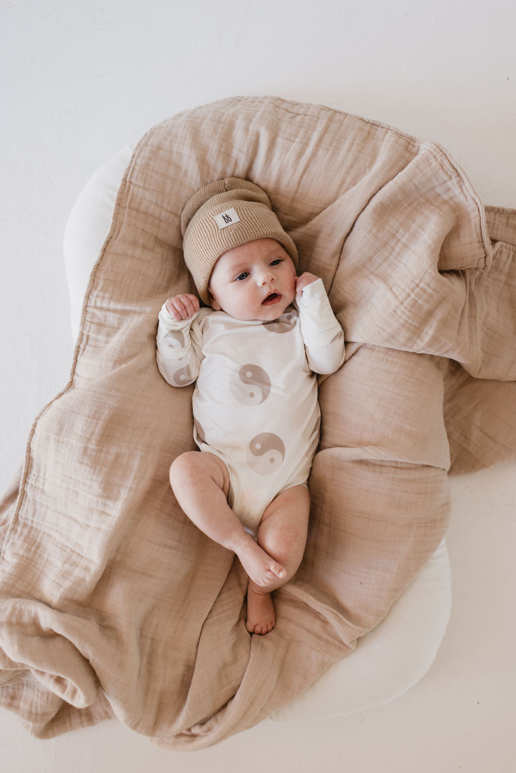 A baby lies on a beige blanket, cozily dressed in forever french baby's Long Sleeve Bamboo Snapsuit | Yin Yang. Made from breathable bamboo fabric, it's gentle and hypo-allergenic. The baby has their arms slightly raised and looks curiously at the camera.