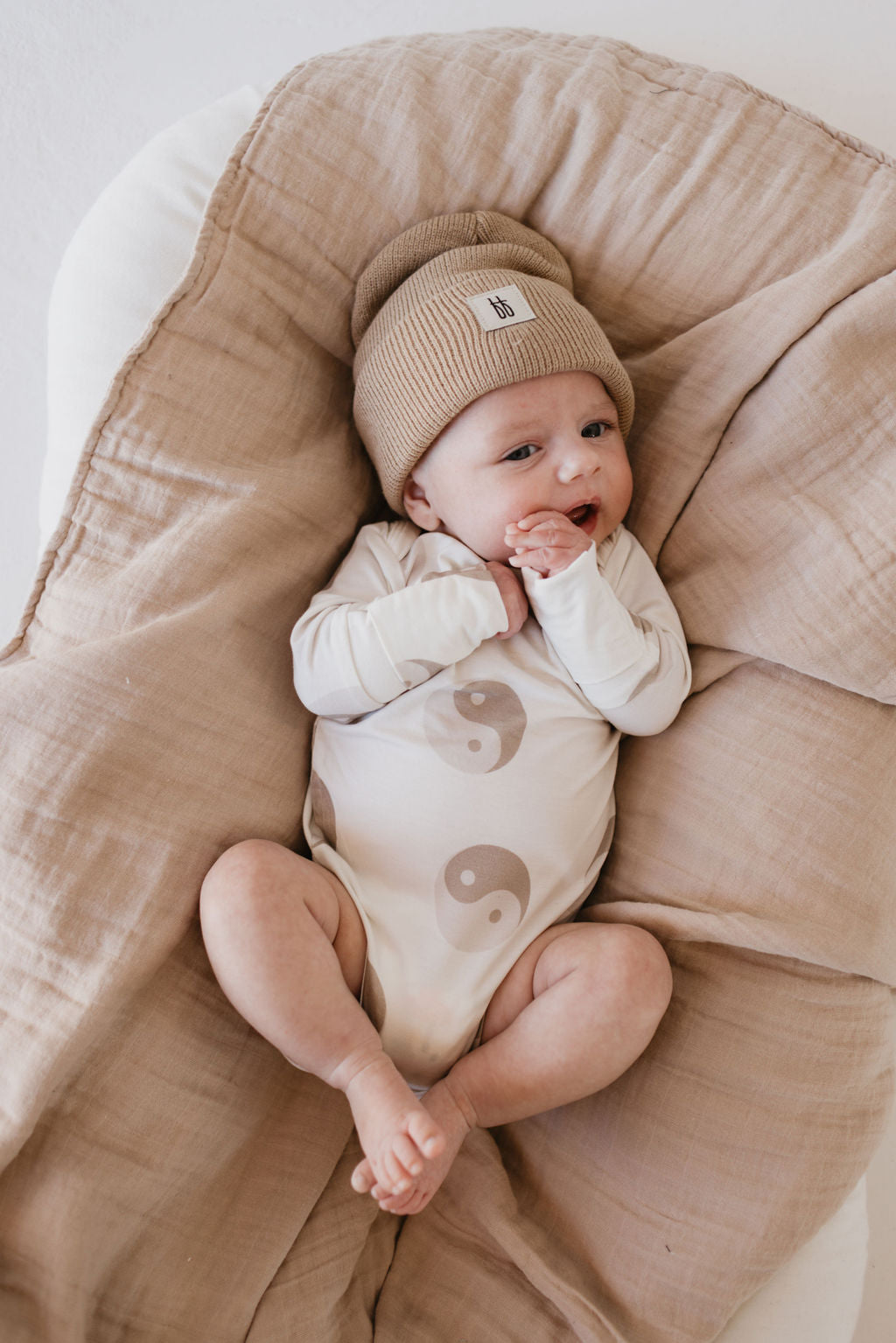 A baby lies content and cozy on a soft tan blanket, wearing a beige beanie and the "Long Sleeve Bamboo Snapsuit | Yin Yang" by forever french baby. Crafted from breathable bamboo fabric, the hypo-allergenic onesie features yin-yang symbols. The baby's hands rest near its mouth.