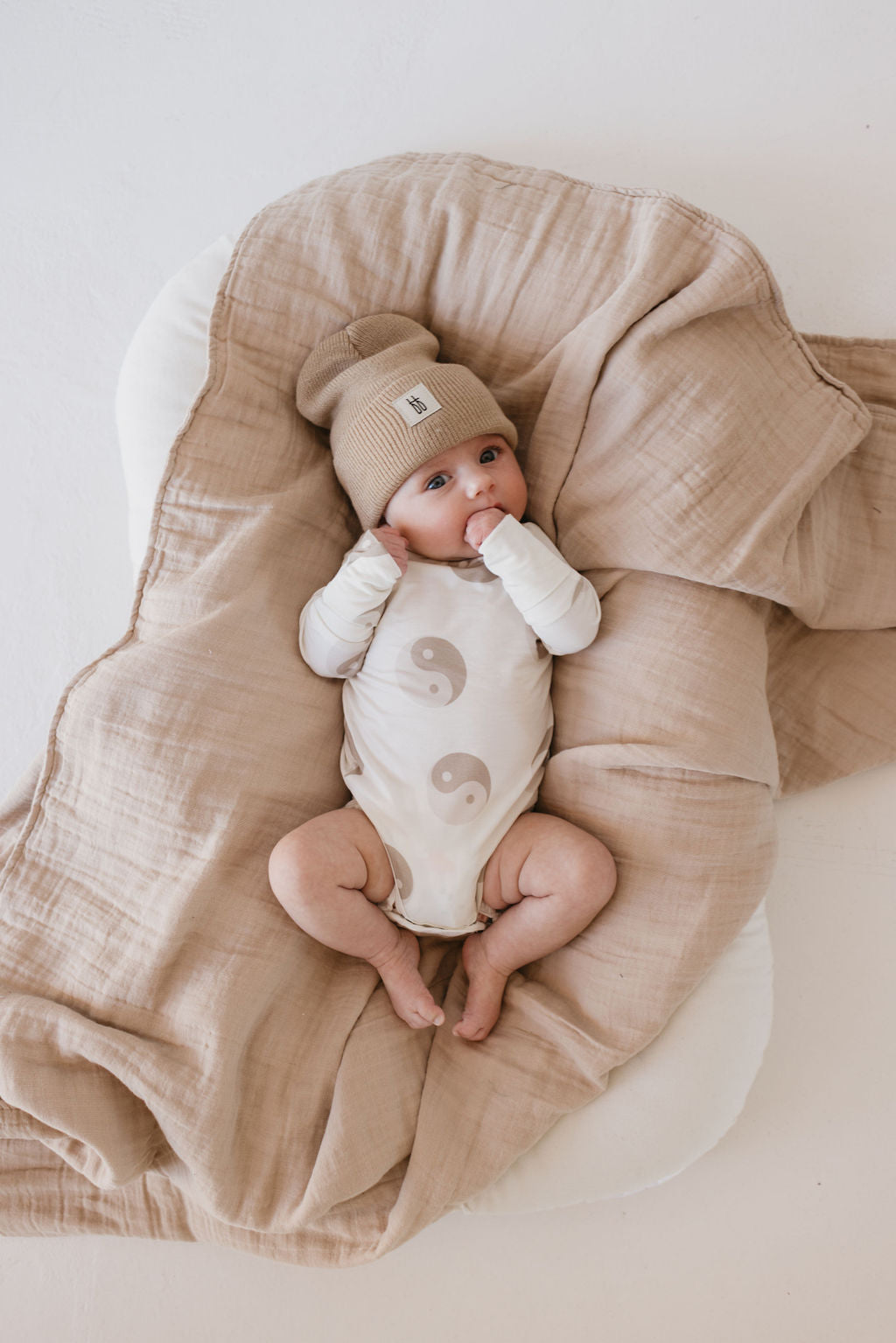 A baby in a beige beanie and the forever french baby Long Sleeve Bamboo Snapsuit | Yin Yang lies on a soft, breathable, hypoallergenic blanket, sucking fingers amid neutral-toned bamboo fabrics.