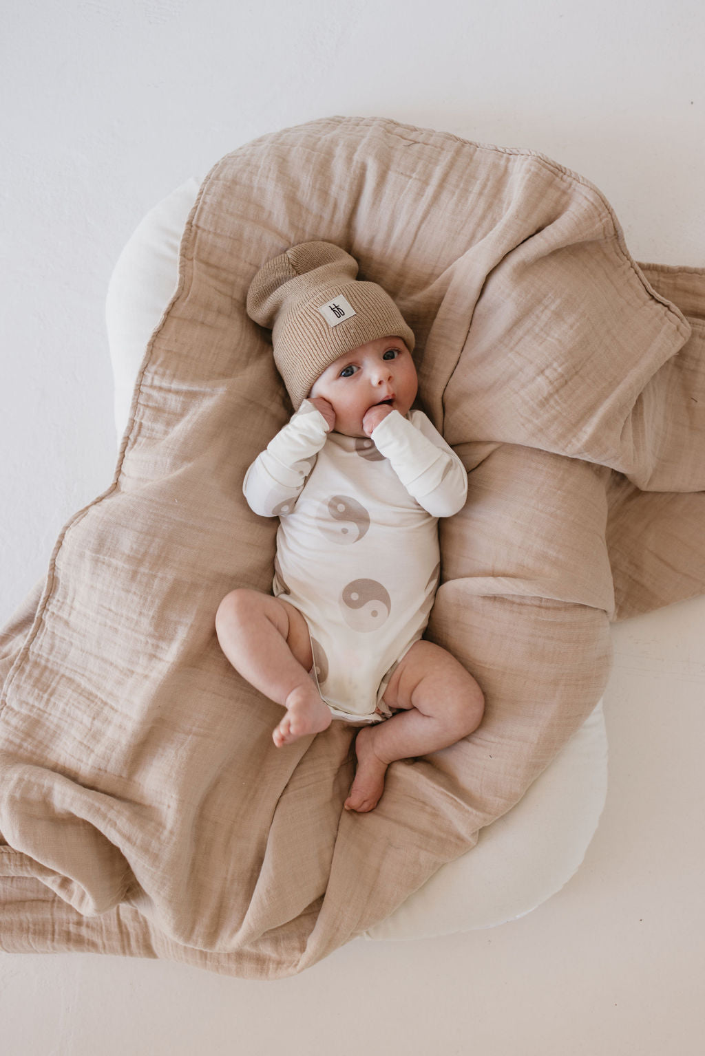 A baby wearing Forever French Baby's Long Sleeve Bamboo Snapsuit in a yin-yang design rests on a soft, hypo-allergenic bamboo blanket. The breathable material envelops the infant as they bring their hands to their mouth and gaze upward.