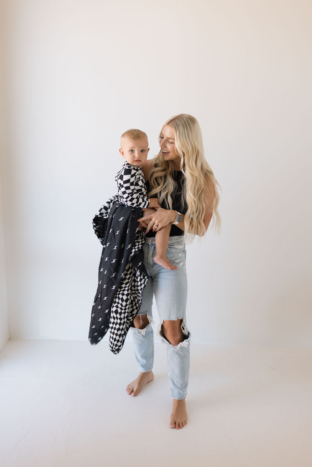 A woman with long blonde hair cradles a young child wrapped in a forever french baby Muslin Quilt featuring Midnight Lightning Bolt and Black + White Wavy Checkerboard designs. She is wearing a black top and ripped jeans, standing barefoot in a minimalist room with a white background.