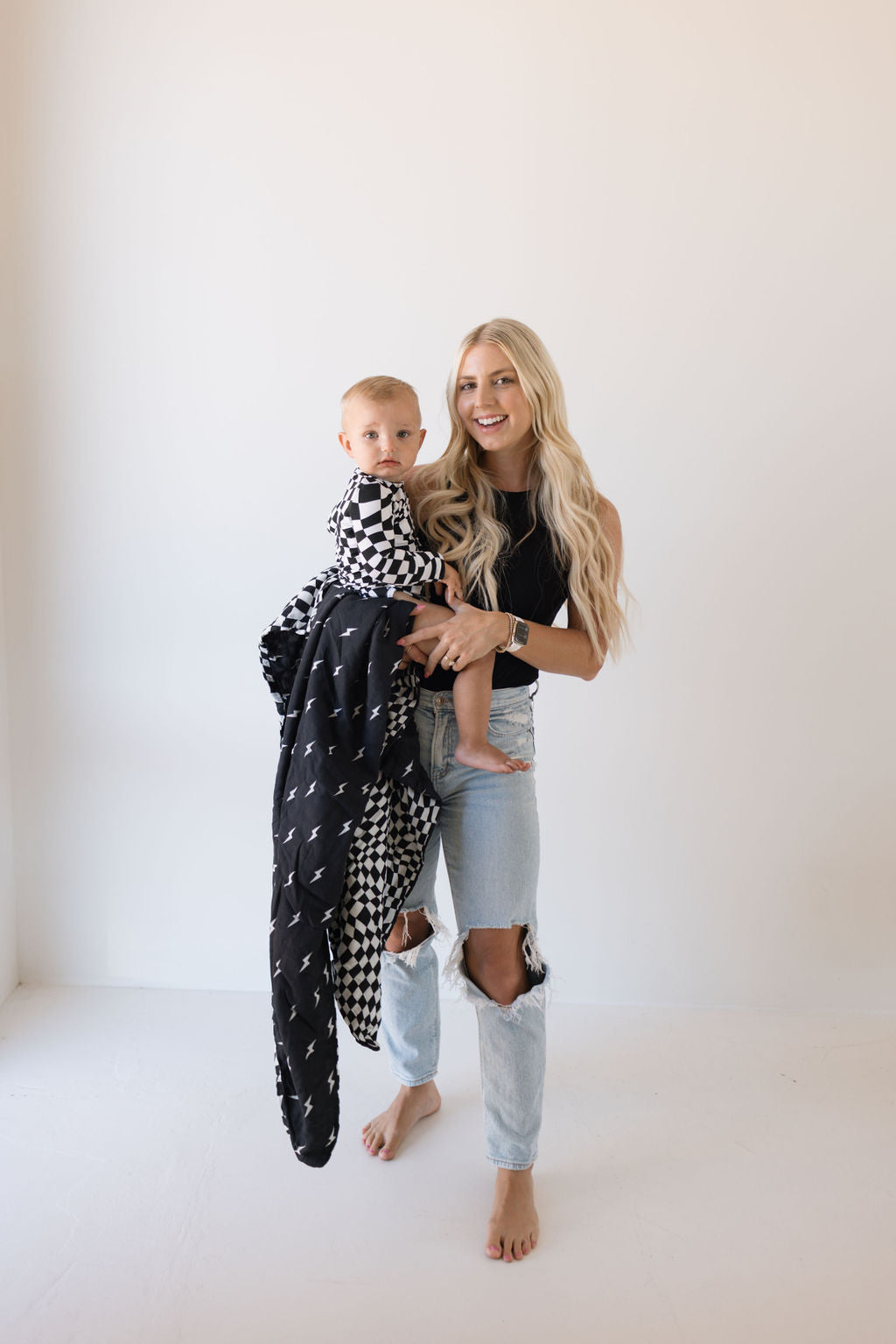 A woman with long blonde hair, clad in a black sleeveless top and ripped jeans, cradles a baby wearing an outfit that mirrors the "Black + White Wavy Checkerboard" design of cozy nursery bedding by forever french baby. Standing barefoot against a plain white backdrop, they exude the warmth reminiscent of the "Muslin Quilt | Midnight Lightning Bolt & Black + White Wavy Checkerboard.