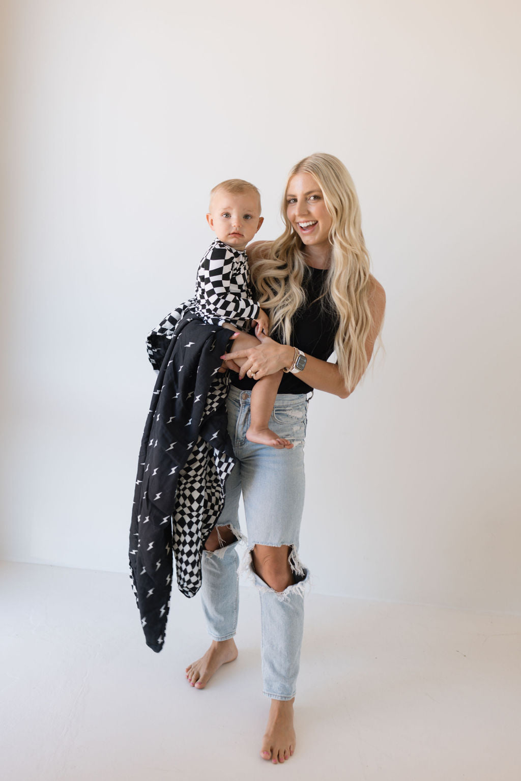 A woman with long blonde hair smiles while holding a child dressed in black and white wavy checkerboard apparel, gently wrapped in a *forever french baby* Muslin Quilt featuring Midnight Lightning Bolt. She is wearing a black top and ripped jeans, standing in a minimalist, light-colored room.